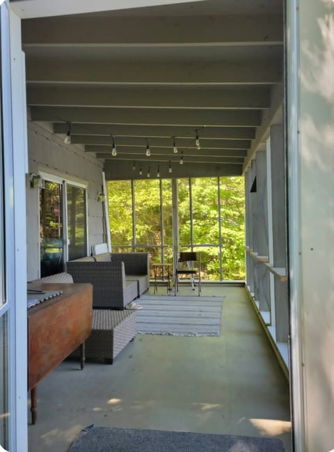 Before: Modern sunroom with exposed beam ceiling, wicker seating, and floor-to-ceiling windows overlooking green foliage