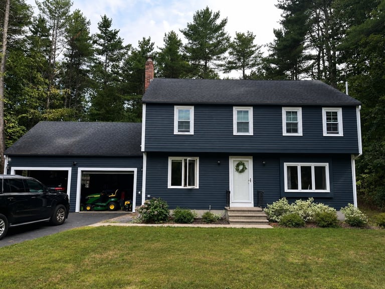 Two-story blue and white house with attached garage, white trim, and landscaped lawn surrounded by evergreen trees