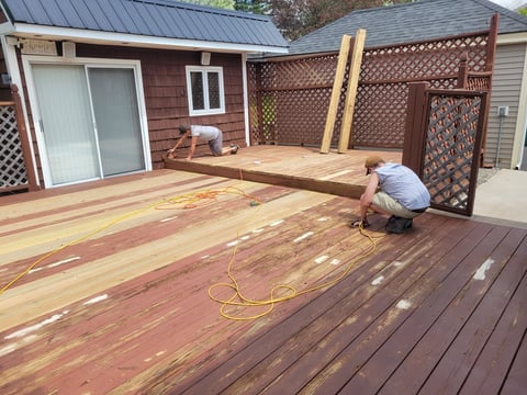 Before: Worker sanding wooden deck during renovation with metal roof cabin and lattice fence in background