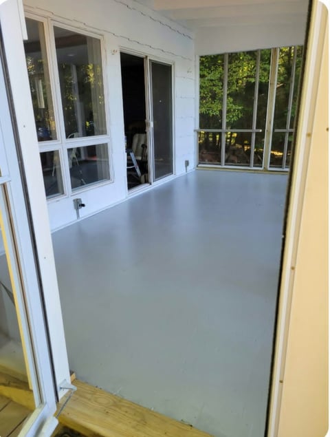 After: Interior view of a covered porch or sunroom with white-framed windows and glass doors overlooking green foliage outside