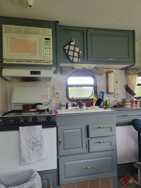 After: Compact RV kitchen with green cabinets, stovetop, microwave, window, and counter space with various items and decor
