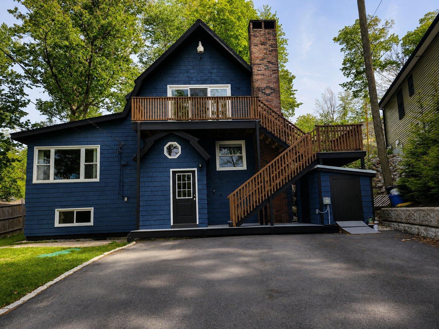 Blue two-story house with brick chimney, wooden deck stairs, driveway, and surrounding green trees