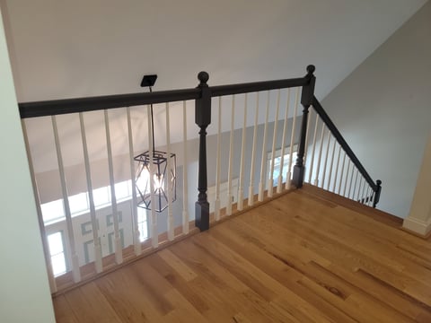 After: Wooden loft landing with black metal stair railing and vertical spindles overlooking lower level with natural light