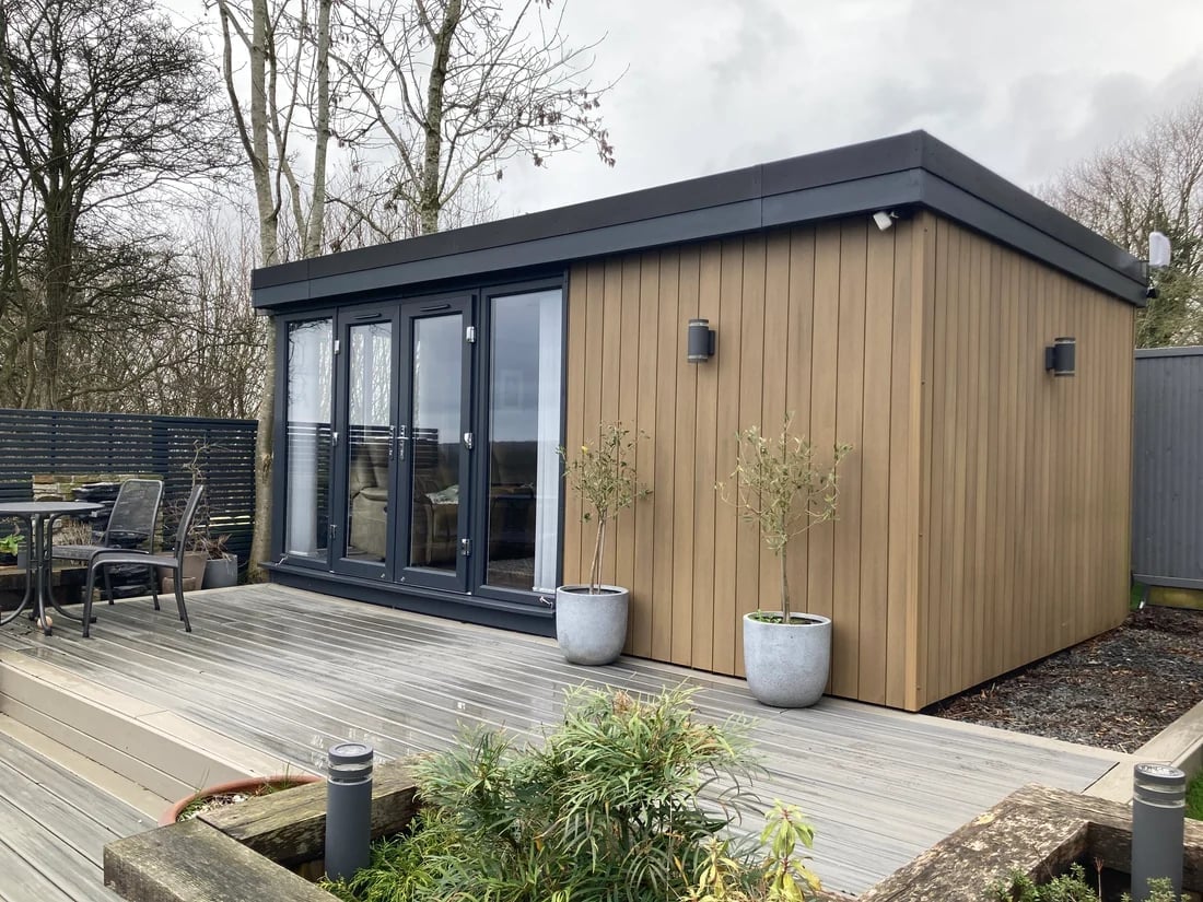Modern garden studio with black frame and timber cladding, positioned on wooden deck with surrounding landscaping
