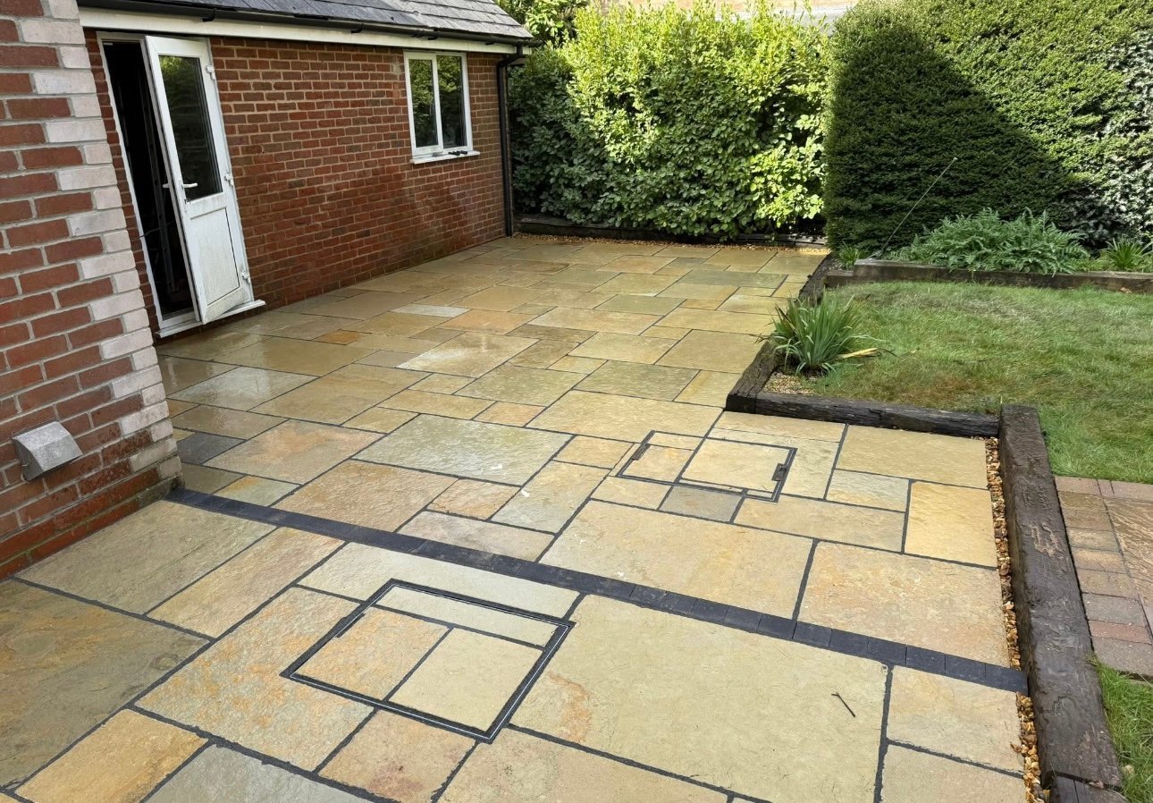 Brick home with a tiled patio area featuring geometric stone patterns, bordered by green hedges and lawn