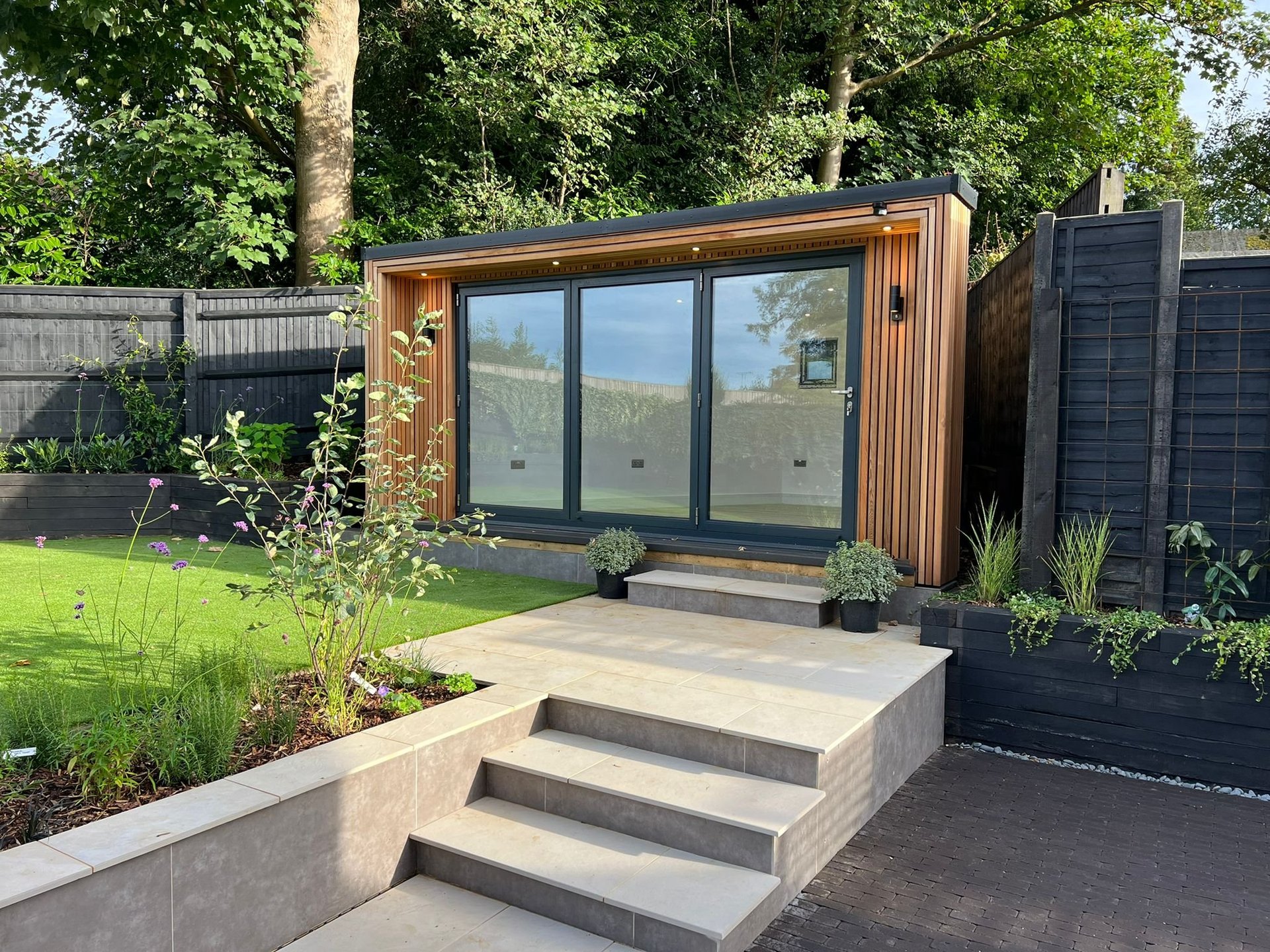 Modern garden studio with wooden cladding and large windows, concrete steps leading to entrance, surrounded by landscaped garden and black fencing