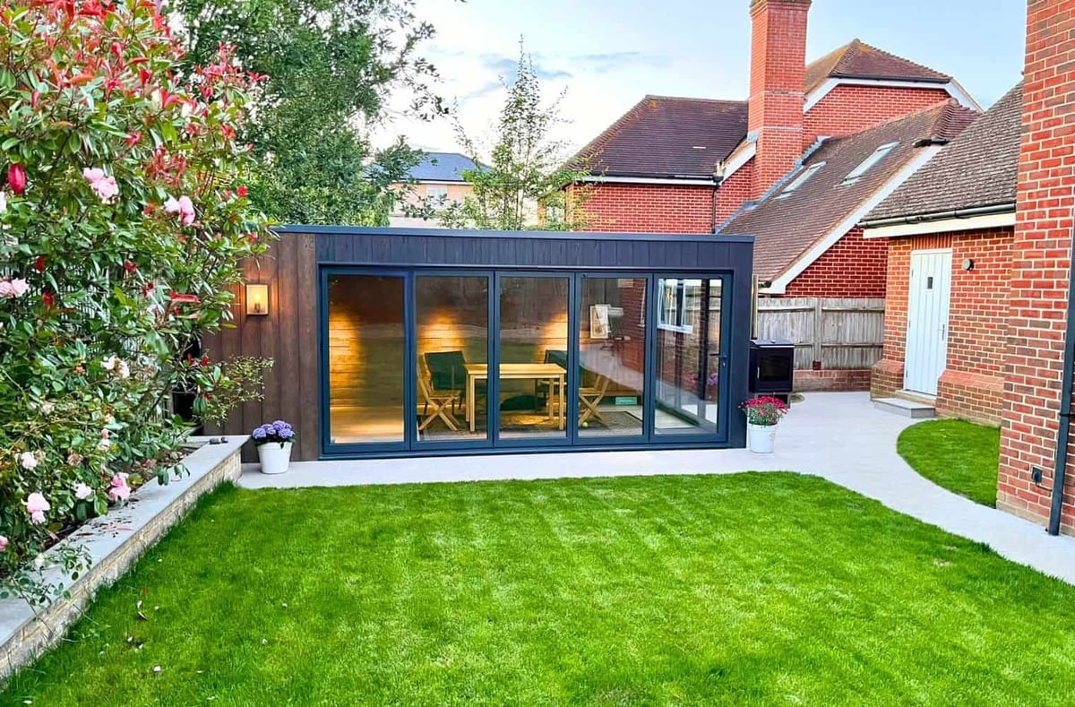 Modern black garden room with glass doors and windows in a brick home backyard with green lawn and flowering vines
