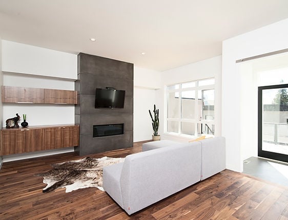 Modern living room with white sectional sofa, dark concrete fireplace with TV, wooden flooring, and large windows overlooking outdoor area