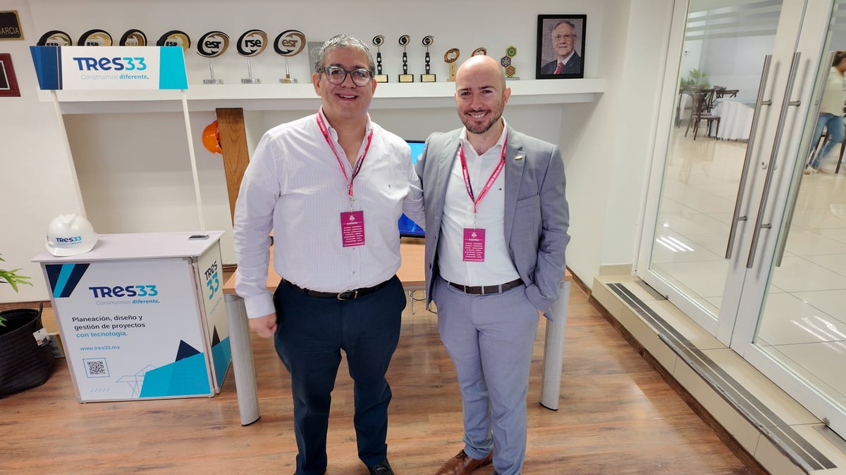Two men wearing glasses and event badges stand together in a modern office with TRES33 branding visible on shelves and displays