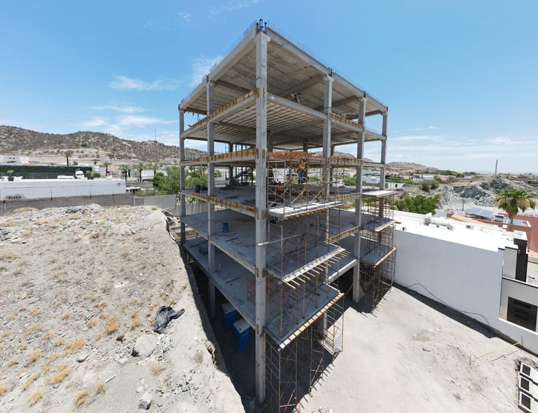 Multi-story concrete building under construction on sandy terrain with mountains and sky in background