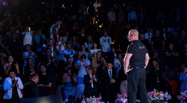 A speaker in black stands on stage addressing a large crowd seated in a dimly lit venue during an event