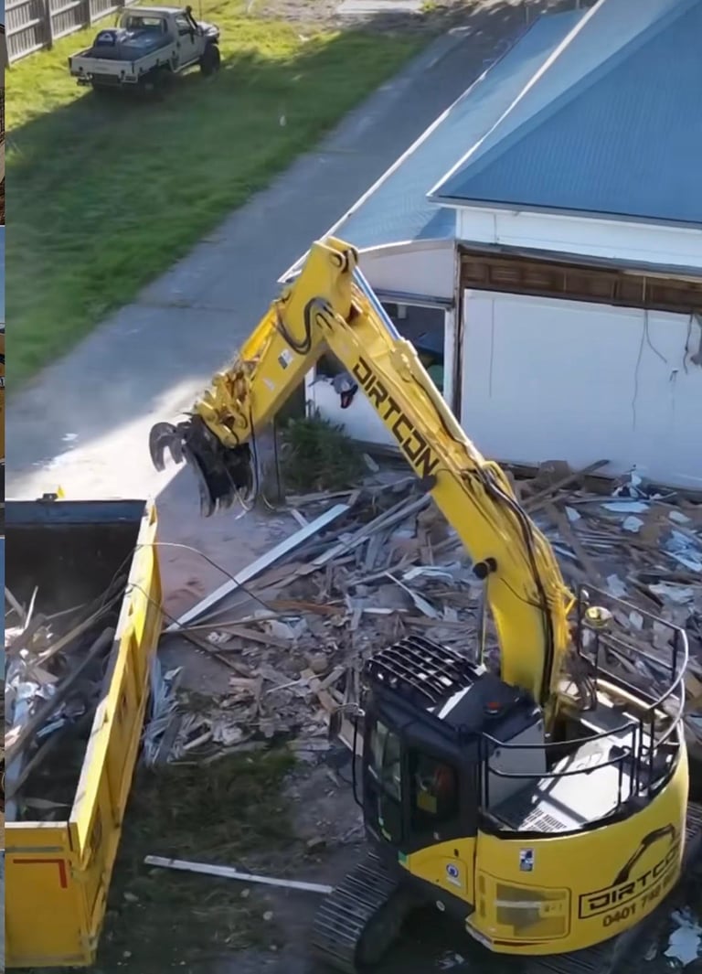 Yellow excavator loading scrap metal into a truck bed at an industrial site with a building in the background