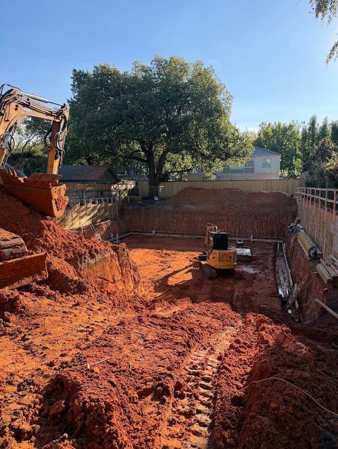 Excavator dumping red clay soil into a fenced construction site with equipment and trees in background