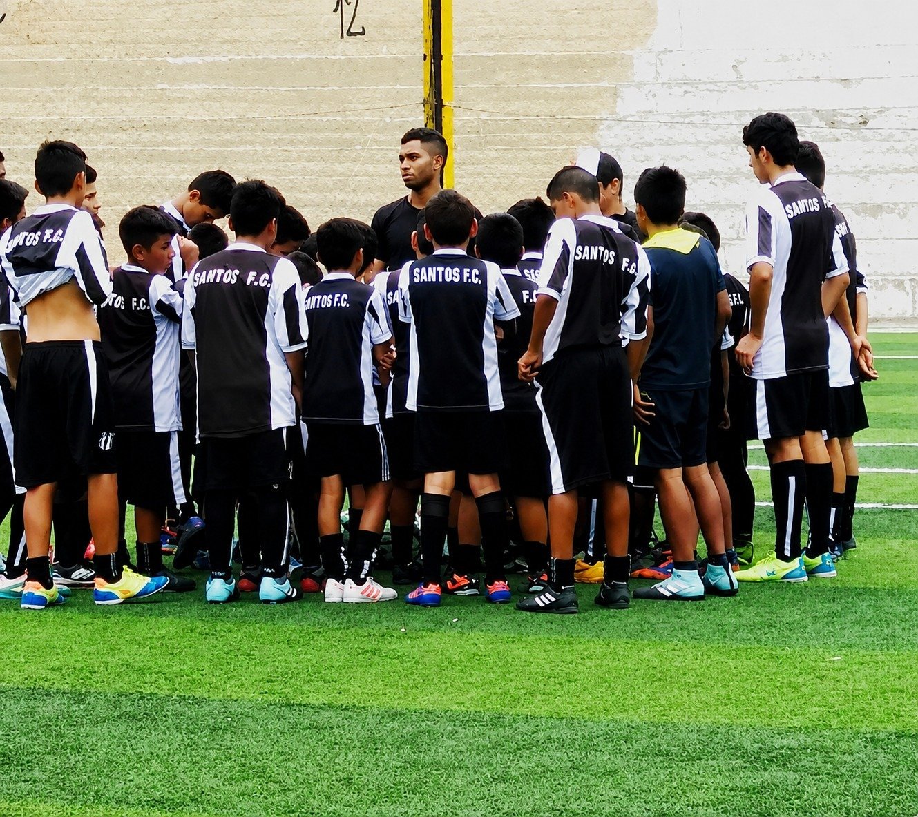 Youth soccer team gathered in a huddle on a grass field wearing black and white Santos FC jerseys during a team discussion.