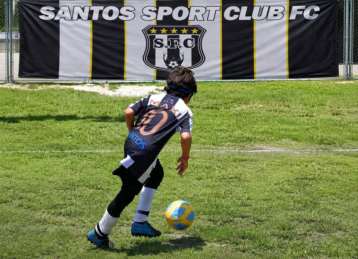 Young soccer player in Santos Sport Club FC jersey dribbling a yellow and blue ball on grass field