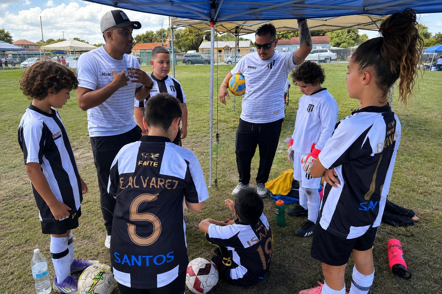 Youth soccer team huddled under blue canopy with coaches during game, players in black and white striped uniforms