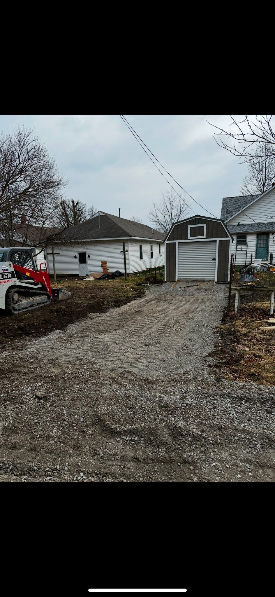 Residential backyard with gravel driveway, white ranch houses, gray barn-style shed, bare trees, and a red skid loader on the left