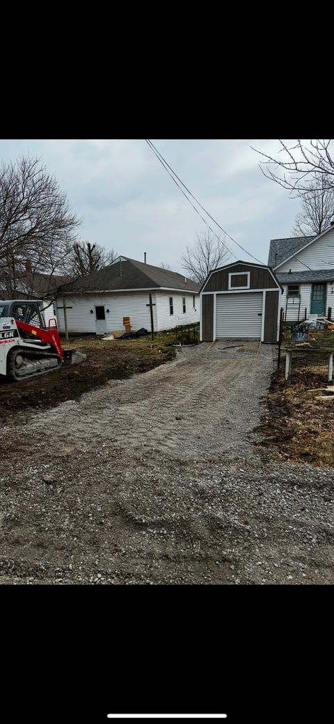 Residential backyard with gravel driveway, white ranch houses, gray barn-style shed, bare trees, and a red skid loader on the left