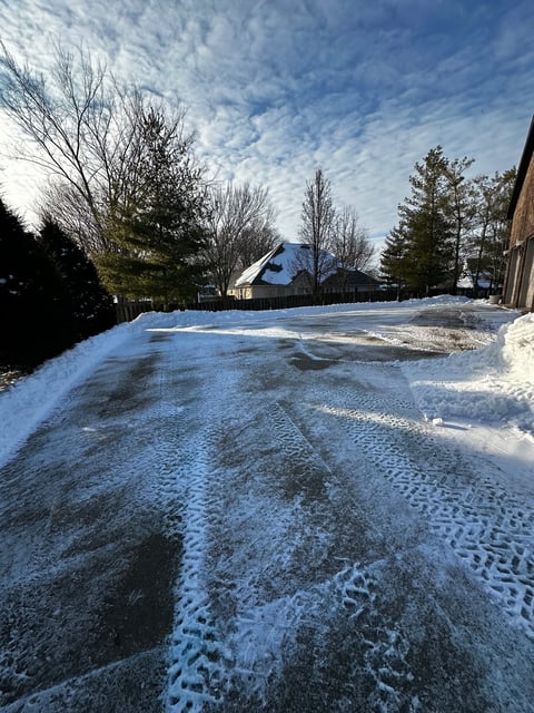 Snow-covered driveway with tire tracks leading to a residential house surrounded by bare winter trees under a blue cloudy sky