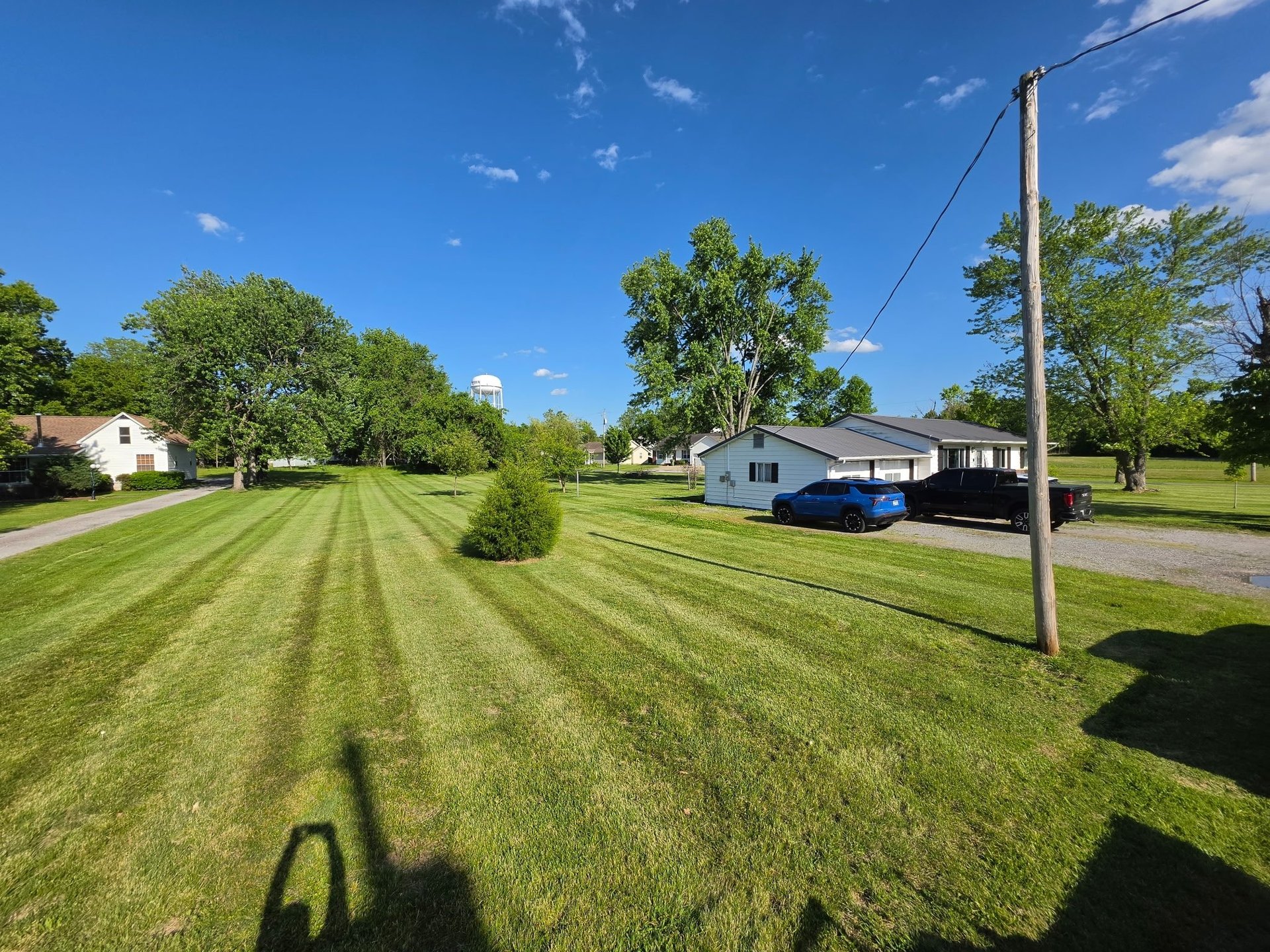 Rural property with manicured lawn, single-story house, utility pole, and green trees under clear blue sky