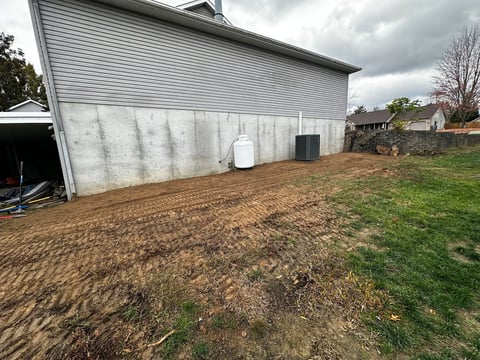 White metal-sided barn with tilled ground and grass yard, cloudy sky, residential area in background