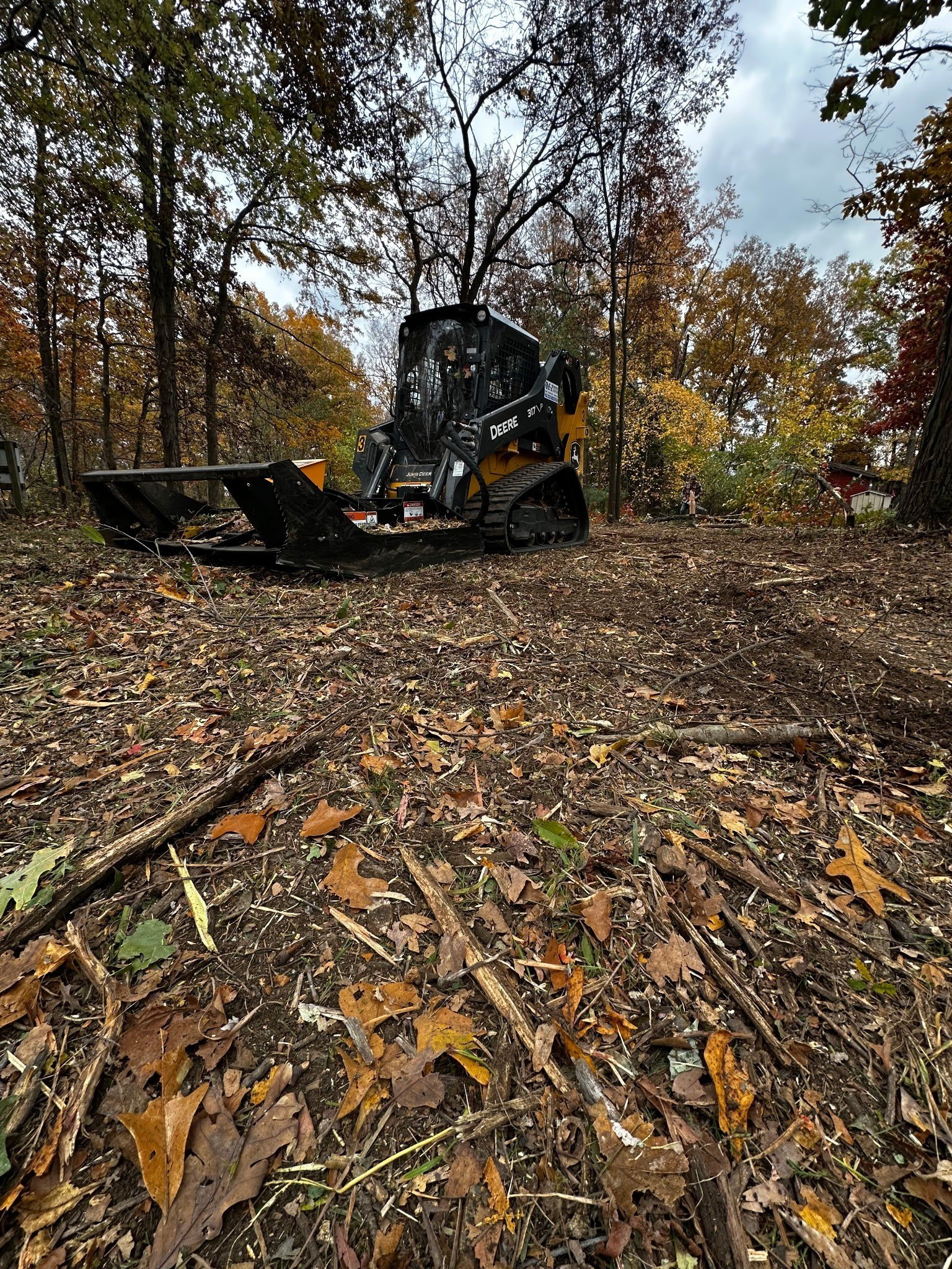 Black skid steer loader clearing fallen leaves and branches in a wooded area during autumn