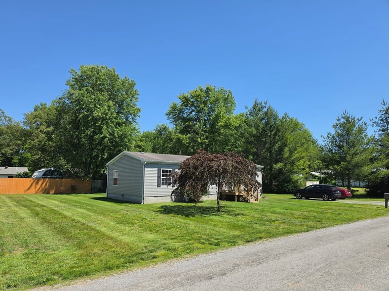 Small gray mobile home on grassy lot with large trees and gravel driveway on clear sunny day