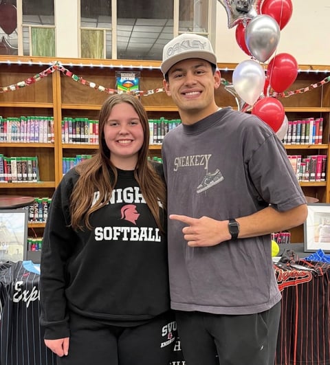 Two students smiling in a library with red and white balloons and a bookshelf backdrop