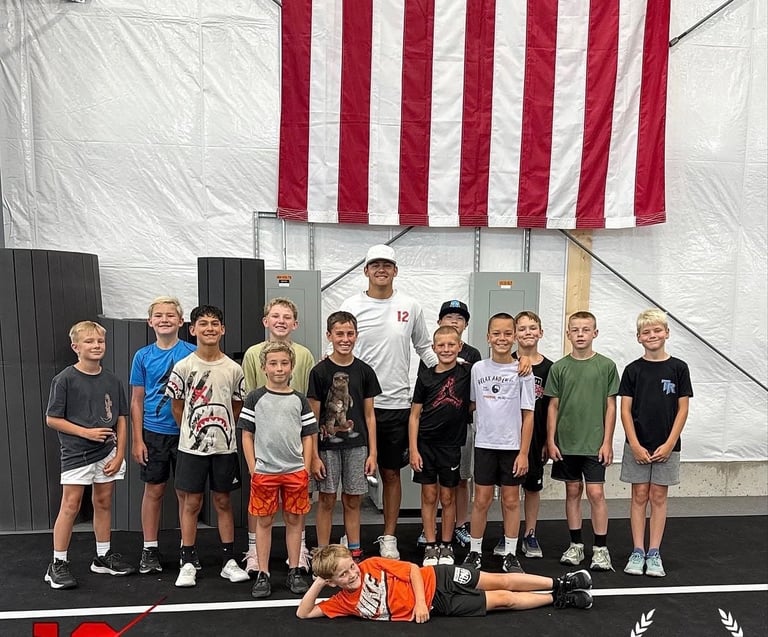 Group of young athletes standing in line under American flag, one child lying on floor in front of gym setting