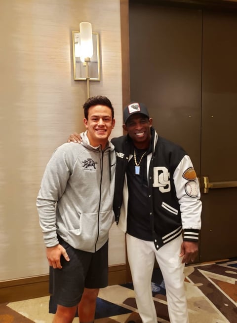 Two men smiling and posing together indoors, one wearing a gray hoodie and the other wearing a black and white varsity jacket with a white cap