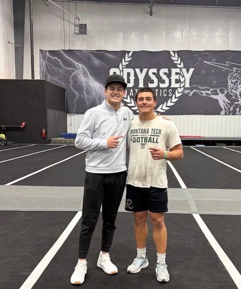 Two male athletes posing together indoors at Odyssey Tactics facility with banner behind them
