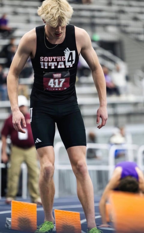 Young male track athlete in black South Utah uniform with race bib 417 standing on indoor track with orange blocks
