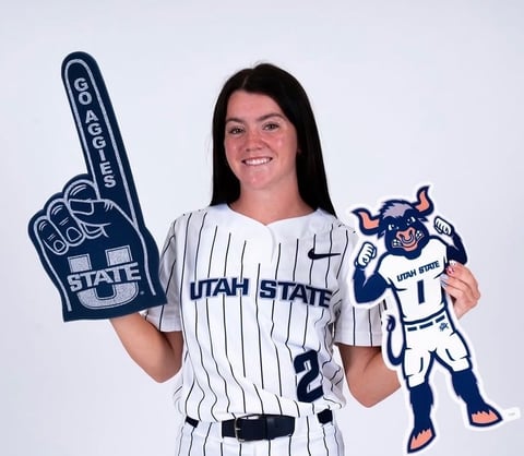 Woman in Utah State baseball uniform holding foam finger and mascot cutout