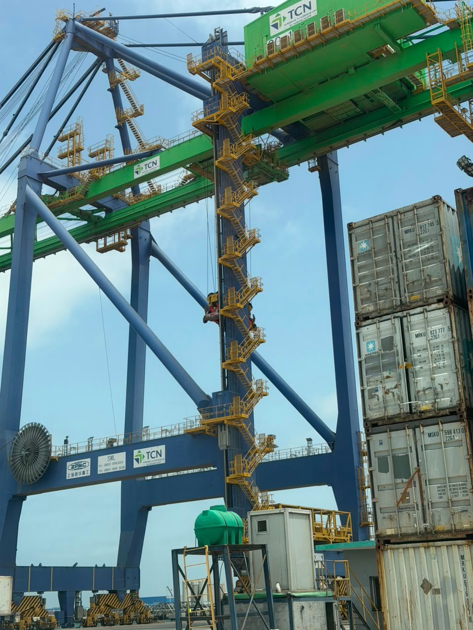 Large green and blue container crane with yellow spreader bars loading cargo containers at a shipping port