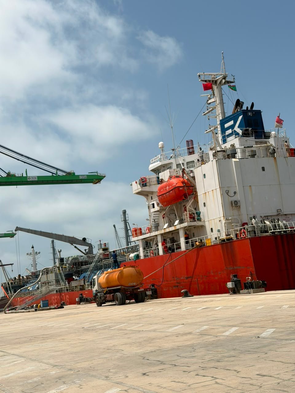 Red and white oil tanker ship docked at port with cargo equipment and other vessels visible in background