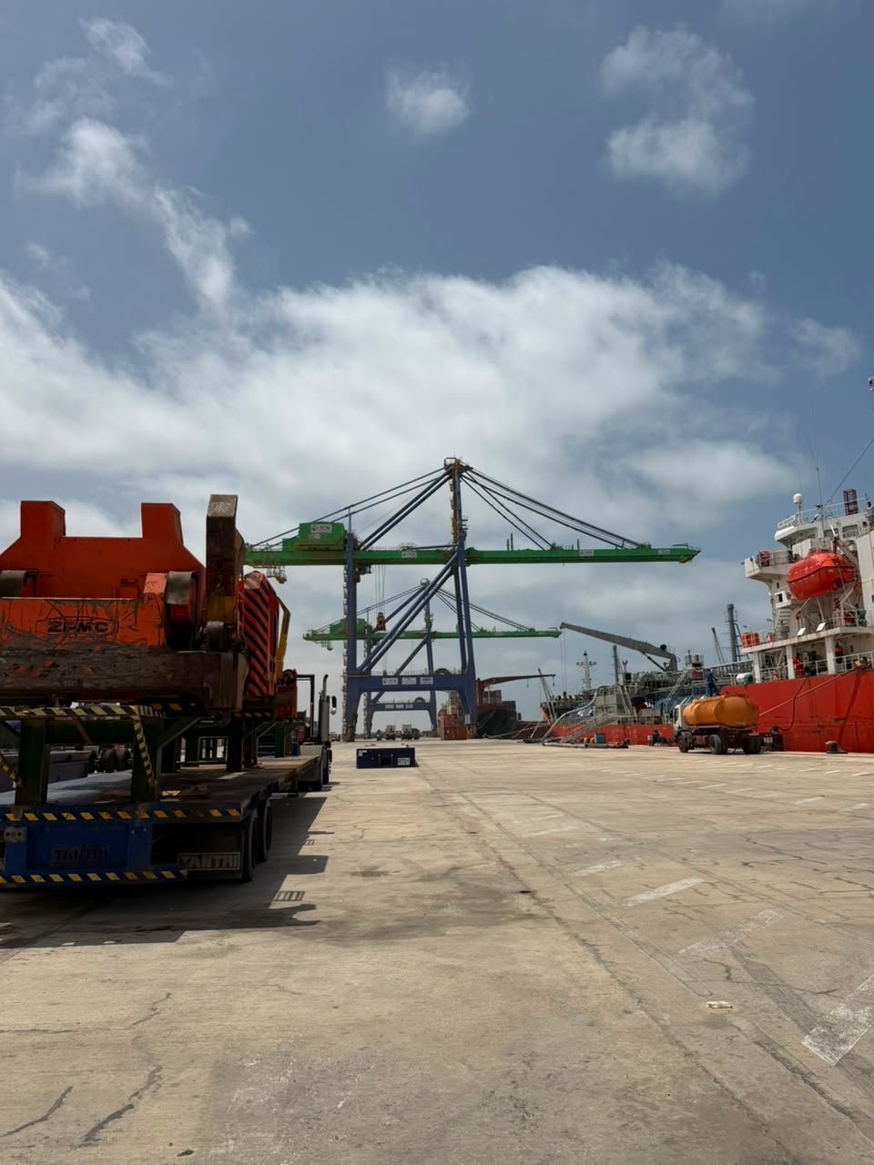 Red cargo truck at industrial port with container gantry cranes and cargo ship under blue cloudy sky