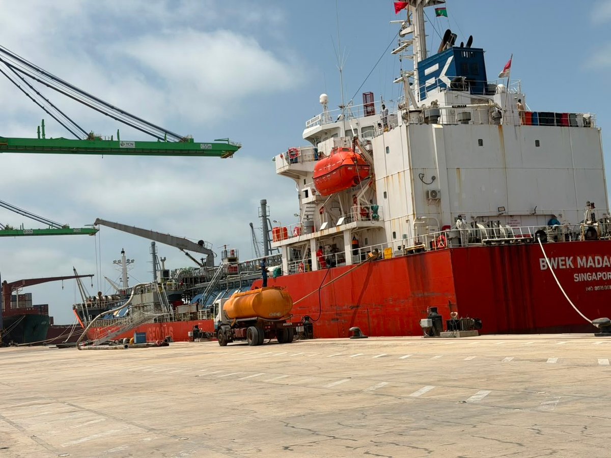 Red and white cargo ship docked at industrial port with loading equipment and container handling cranes visible