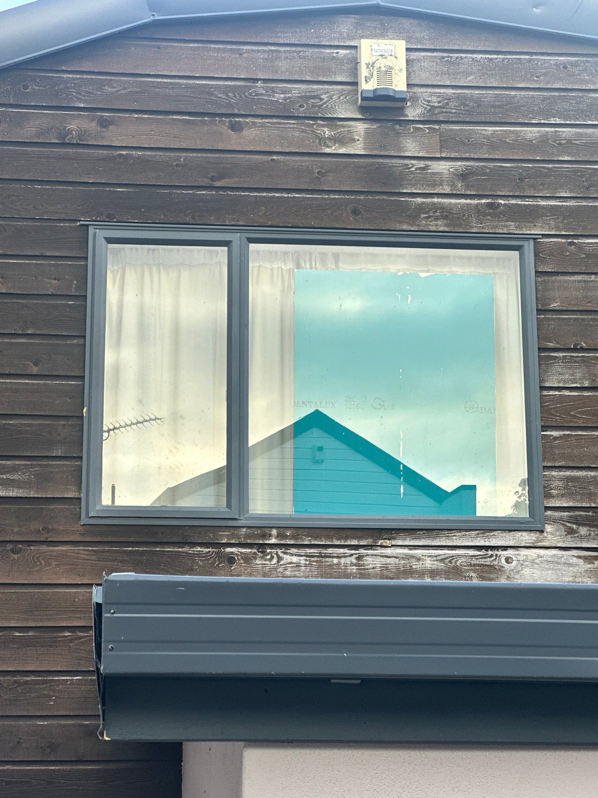 Window in weathered dark wood building with turquoise beach hut visible in reflection and through glass