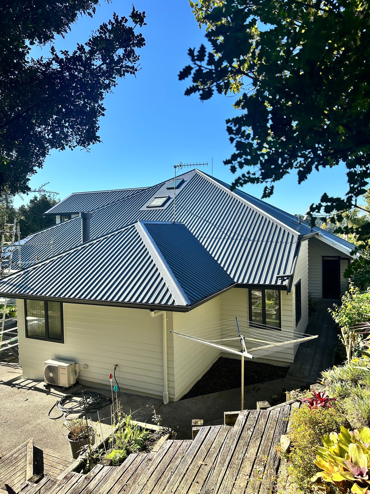 Modern cream and black house with metal roof surrounded by green trees and wooden deck