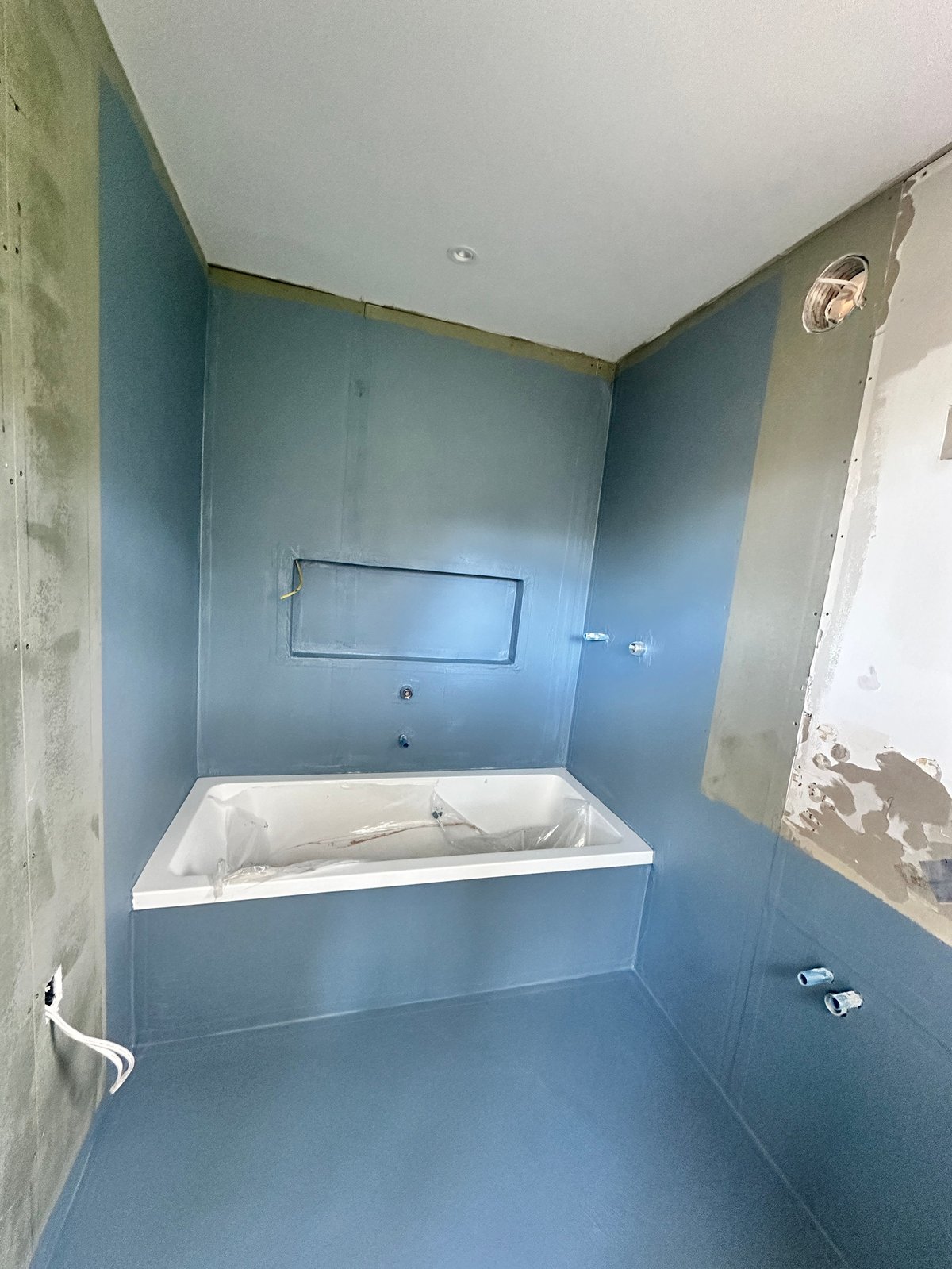 Bathroom under renovation with blue-painted walls, white bathtub, and exposed drywall, incomplete plumbing installation visible