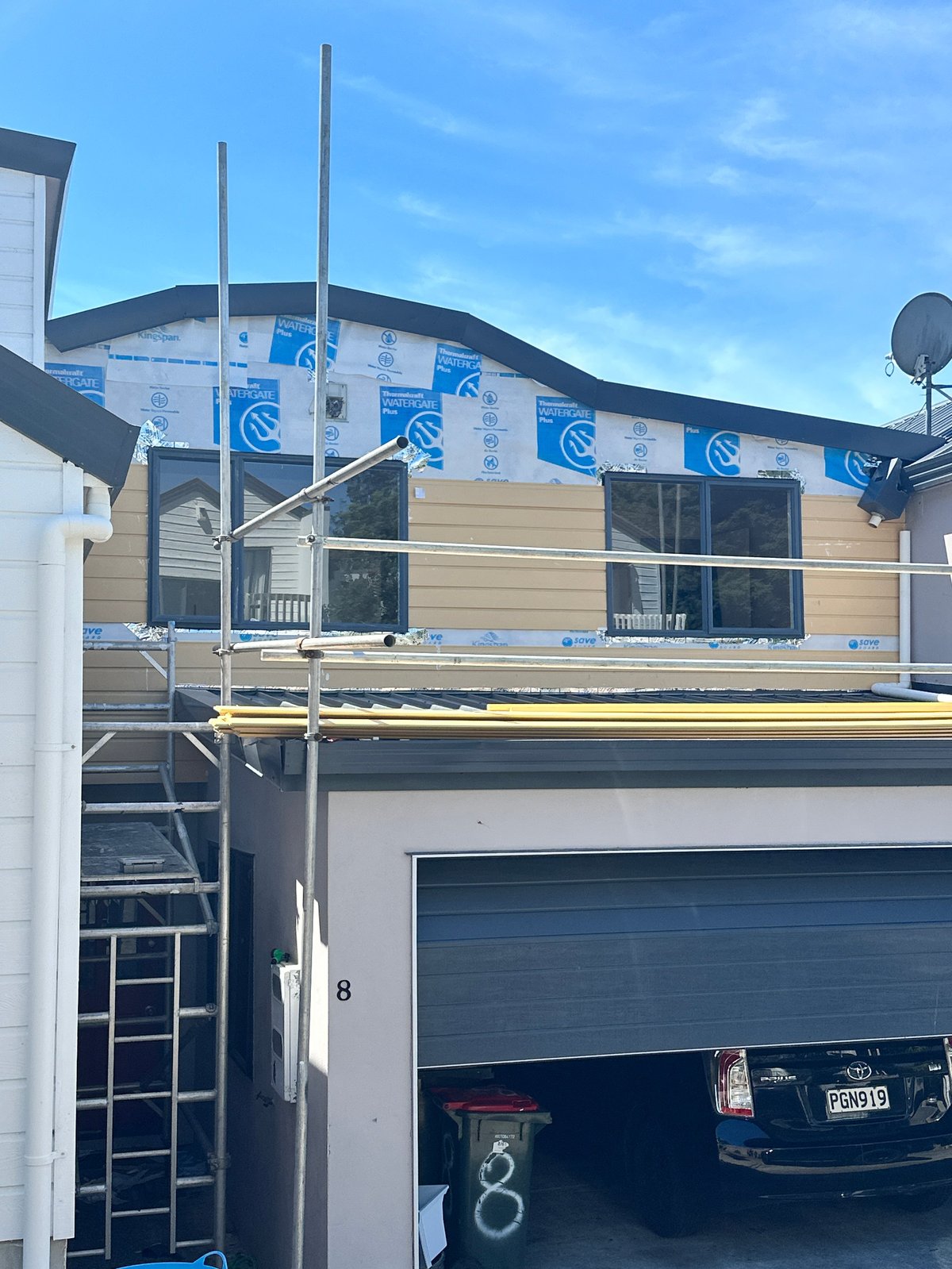 New home under construction with blue protective wrapping, metal scaffolding, garage with car parked inside, clear sky