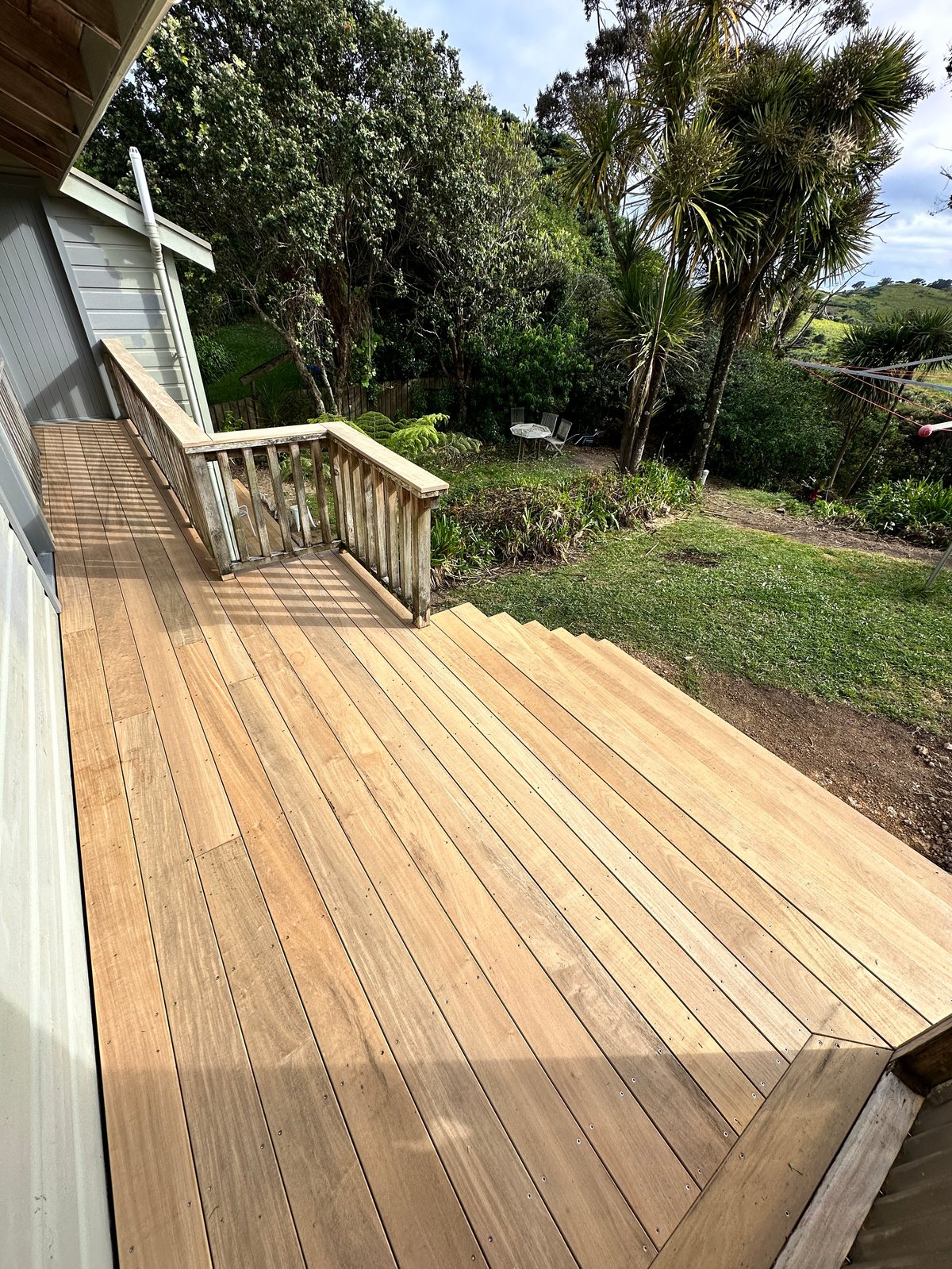 Wooden deck and stairs attached to a white house, overlooking a lush tropical garden with palm trees and green landscape