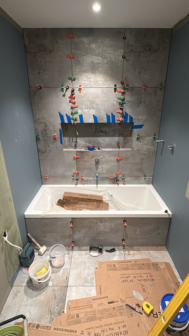 Bathroom renovation in progress showing tiled shower wall with marked fixtures, white tub below, and construction materials on floor