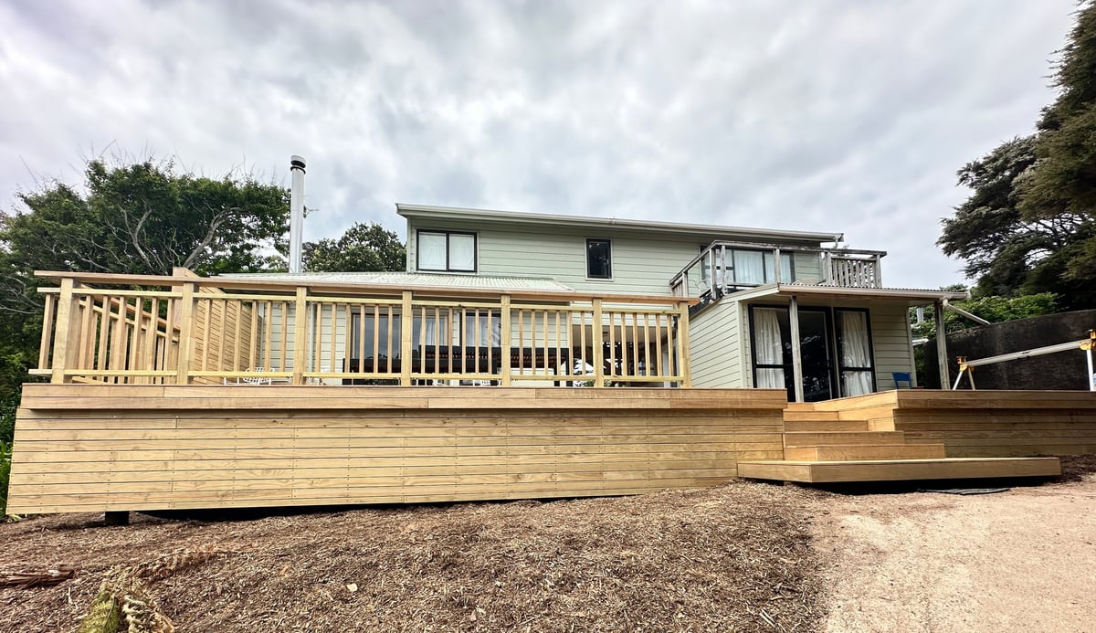 Modern two-story house with light green exterior and large wooden deck overlooking gravel yard on cloudy day