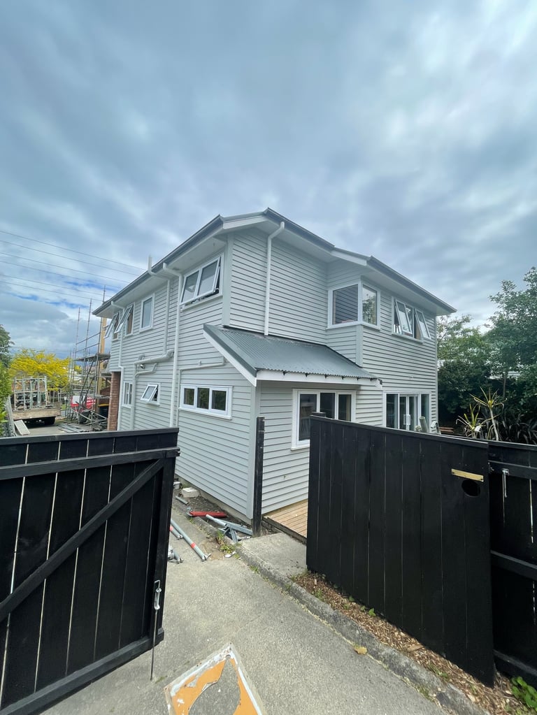 Modern white two-storey Hauraki home after recladding, with black gates and driveway under a cloudy sky