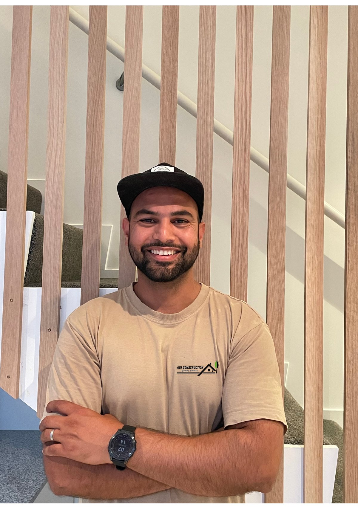 Smiling man wearing a beige t-shirt and black cap, arms crossed, against a wooden slat wall background