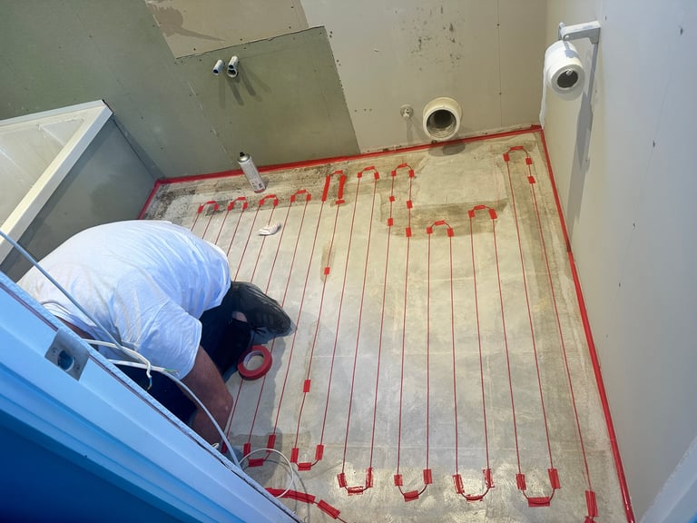 Worker in blue uniform installing red radiant heating tubes on floor with tape outline marking the heating system layout
