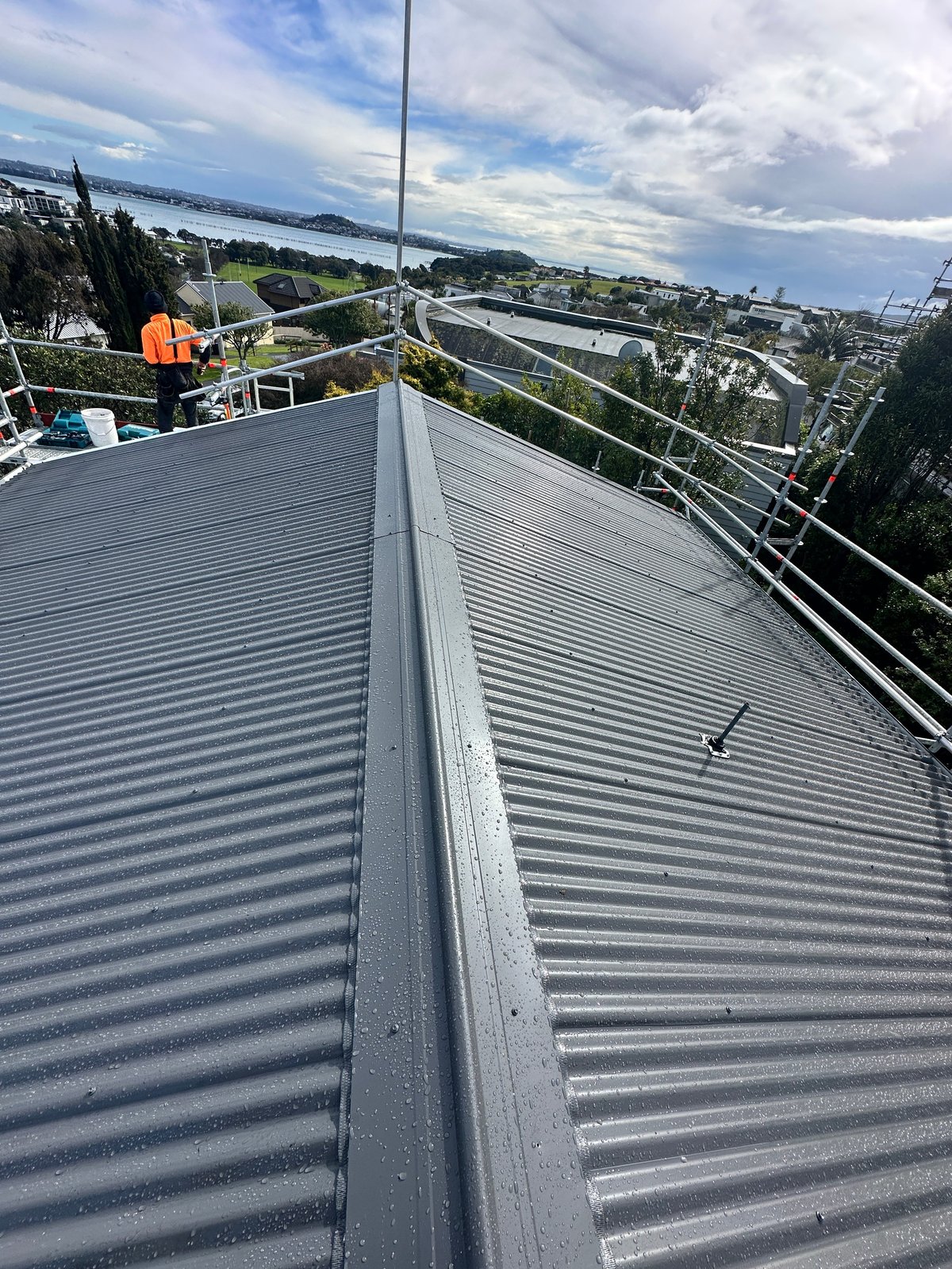 Worker in orange safety gear laying the new corrugated roofing with the harbour and city behind