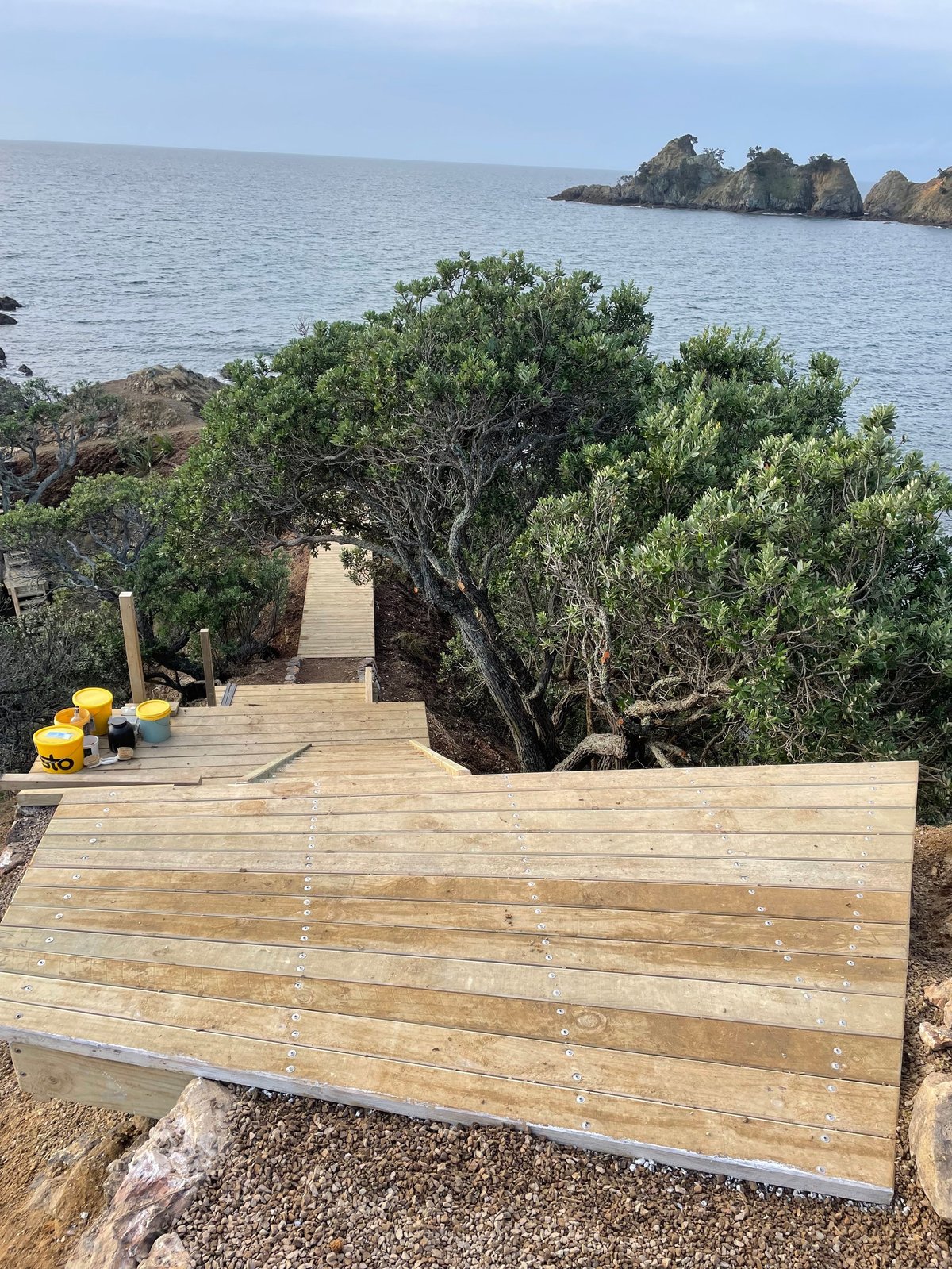 Wooden deck platform overlooking a coastal bay with lush green shrubs, rocky outcrops, and calm blue water