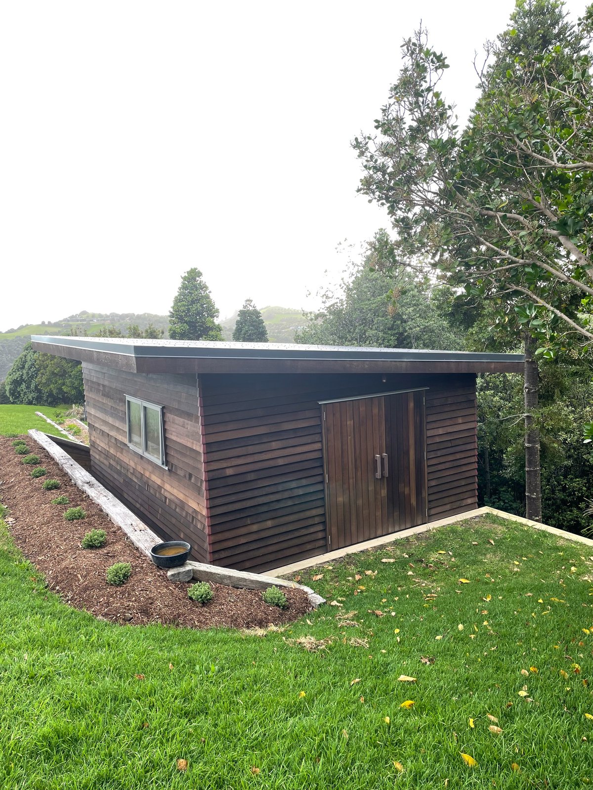 Modern dark cedar cabin with flat roof set in green lawn and trees on a Waiheke Island hillside